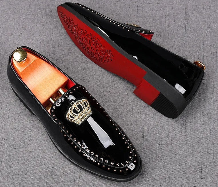 A pair of black patent leather men's loafers with a gold crown embroidery and a red bottom sole, displayed against a grey background.