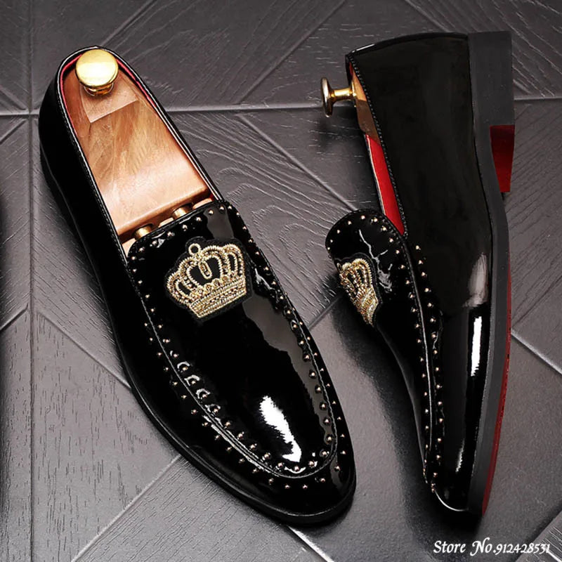 A pair of black patent leather men's loafers with a gold crown embroidery and a red bottom sole, displayed against a grey background.