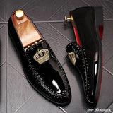 A pair of black patent leather men's loafers with a gold crown embroidery and a red bottom sole, displayed against a grey background.